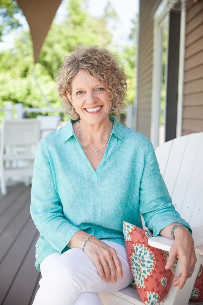 Image of Rhonda. She sits outside on a deck in a white chair next to a coral colored pillow. She is wearing an aqua shirt.